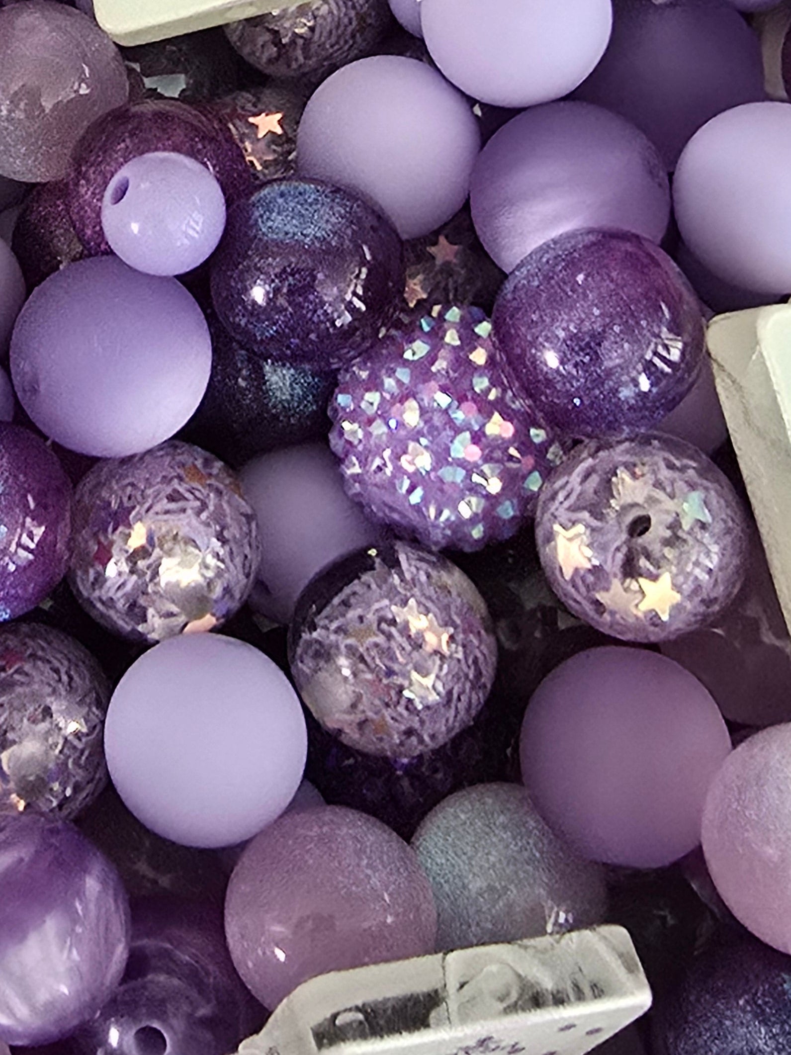 A dense pile of purple and lavender craft beads in various sizes, including smooth matte spheres, glittery round beads, and small white decorative padlocks with purple ribbon bows.