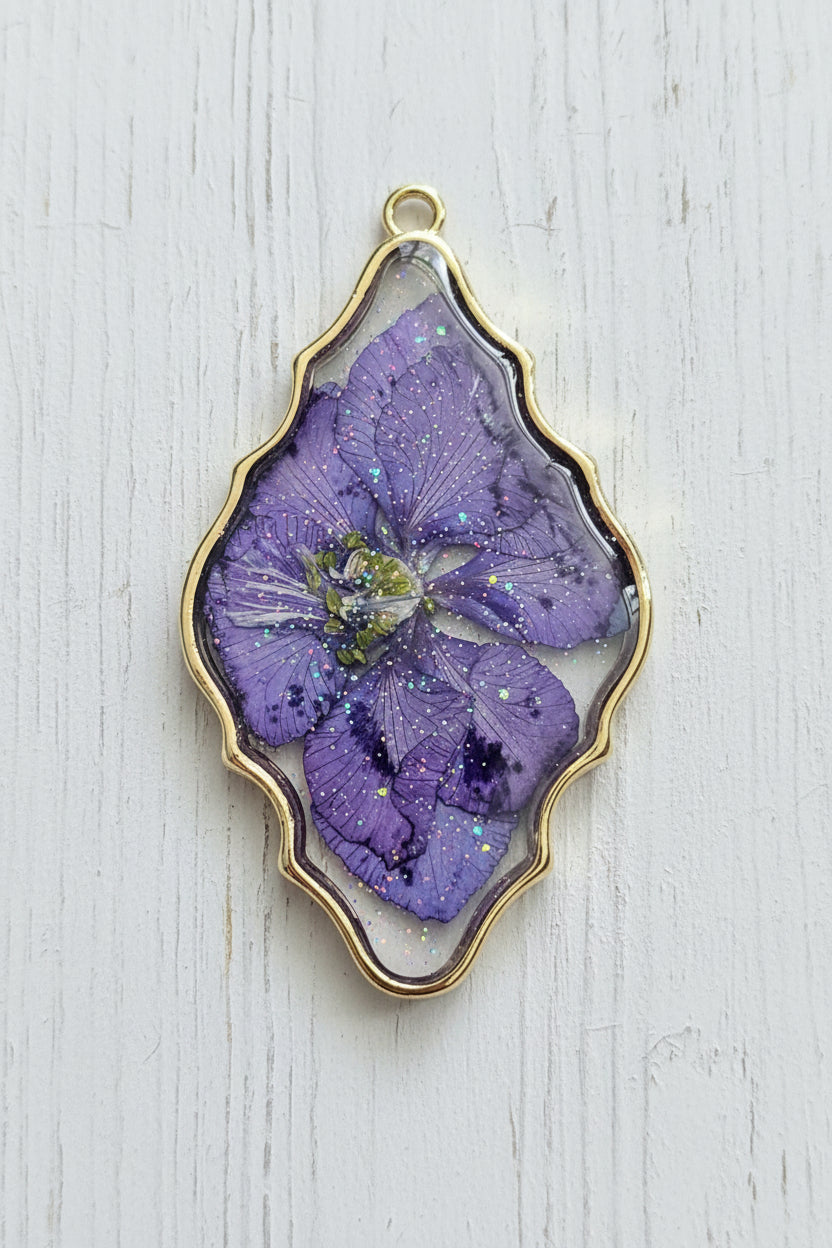 A scalloped oval pendant featuring a preserved purple flower inside a gold-toned metal frame