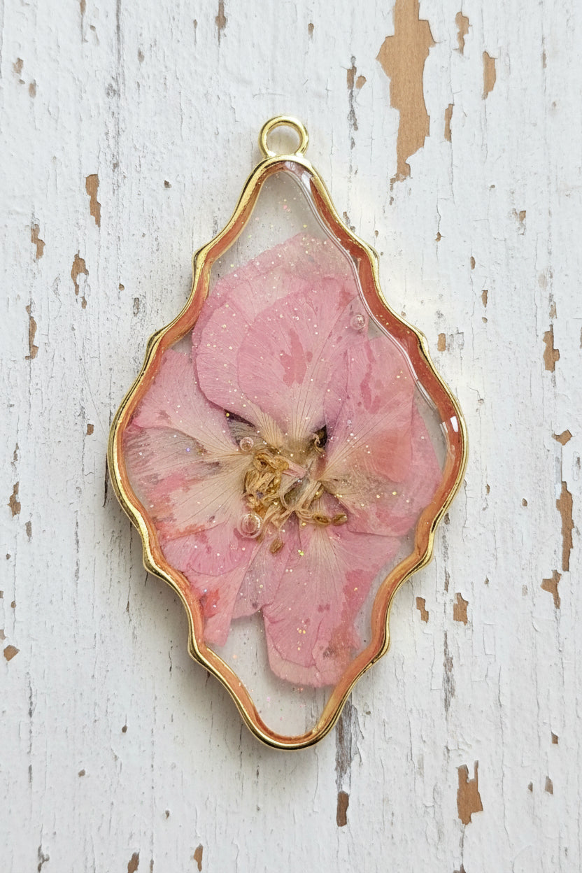 A scalloped oval pendant featuring a preserved pink flower inside a gold-toned metal frame