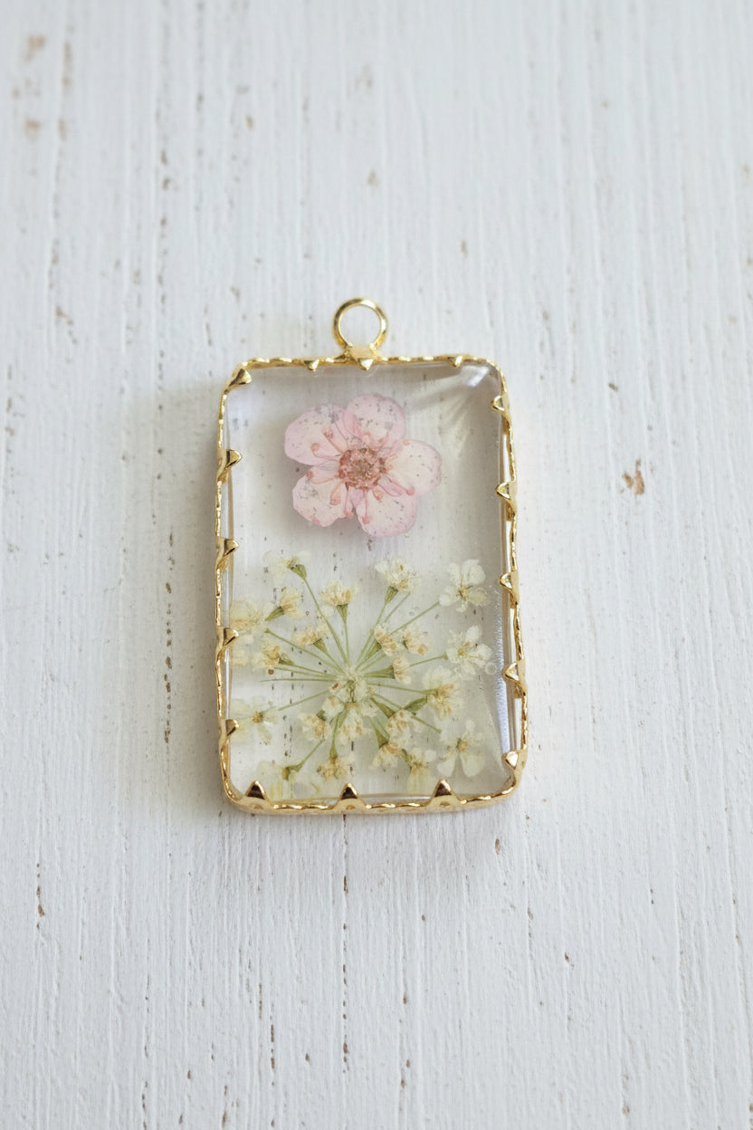 A rectangular clear resin pendant or charm with a gold-tone border, featuring a pressed pink flower and blue dandelion-like elements