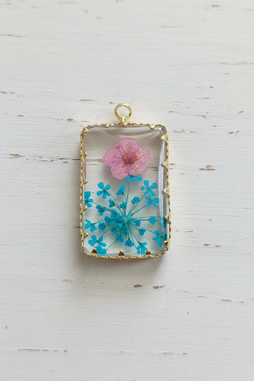 A rectangular clear resin pendant or charm with a gold-tone border, featuring a pressed pink flower and blue dandelion-like elements