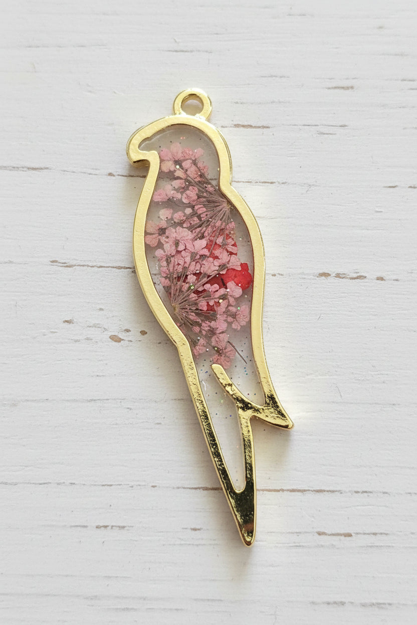 A gold-plated metal bird-shaped pendant or charm with a small hanging loop, filled with dried pink and red flowers