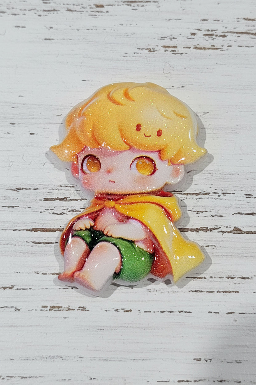 A small glossy acrylic bead of a cute cartoon child with yellow hair, orange eyes, a yellow cape, and green clothing, placed on a white distressed wooden surface.