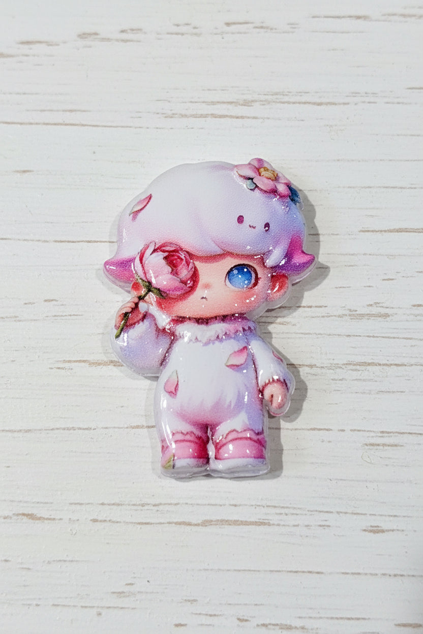 A small glossy character acrylic bead featuring a pastel pink and white doll with a blue eye, floral hair accessory, and pink bow, placed on a whitewashed wood surface.