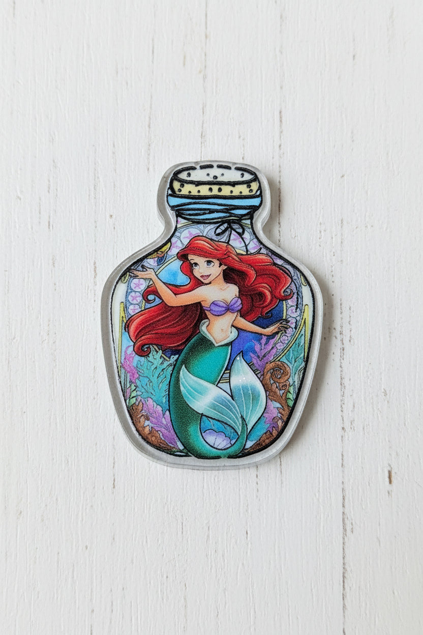 A small decorative bottle-shaped charm or pendant featuring an illustrated mermaid with red hair and a green tail.