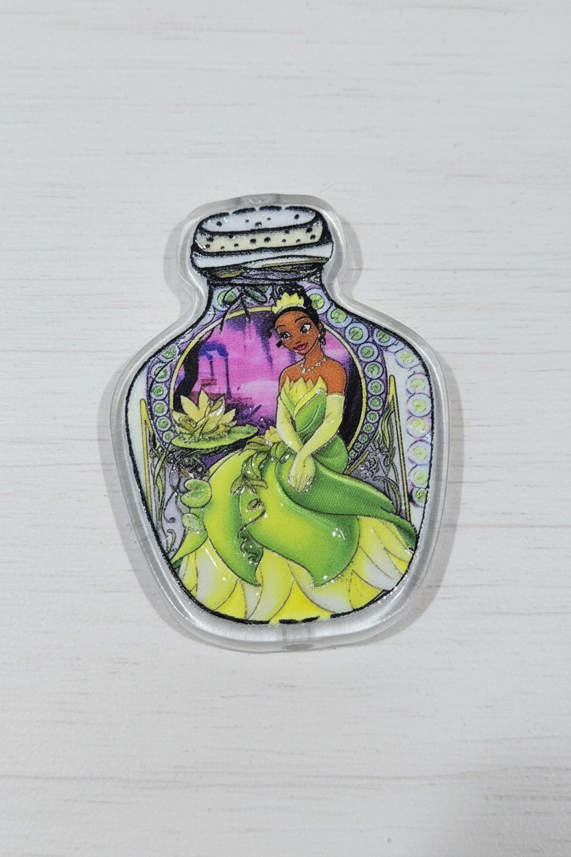 A small clear acrylic bottle-shaped charm or pendant featuring a colorful illustration of a green frog-like character and a purple background.