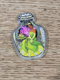 A small clear acrylic bottle-shaped charm or pendant featuring a colorful illustration of a green frog-like character and a purple background.