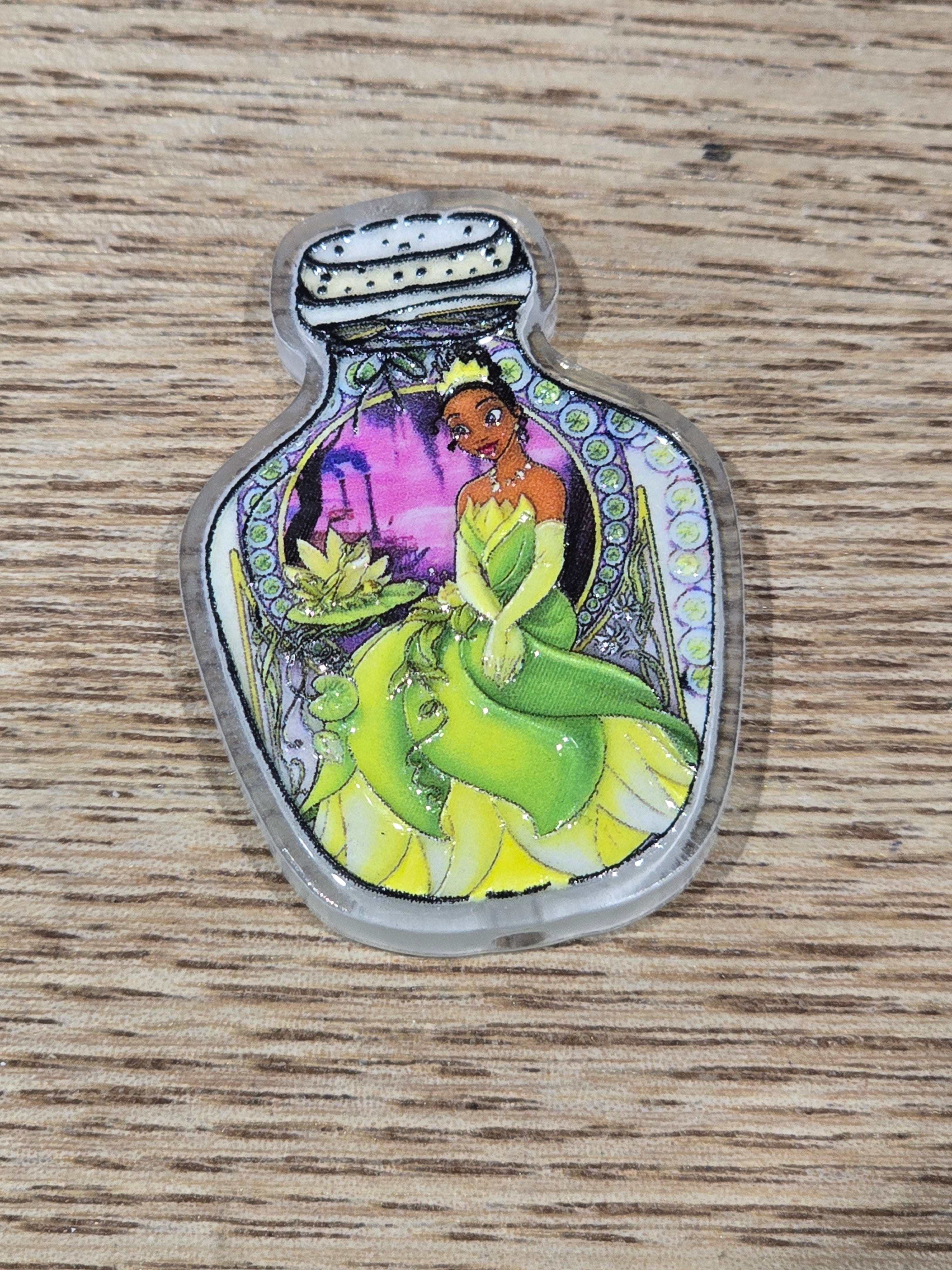 A small clear acrylic bottle-shaped charm or pendant featuring a colorful illustration of a green frog-like character and a purple background.
