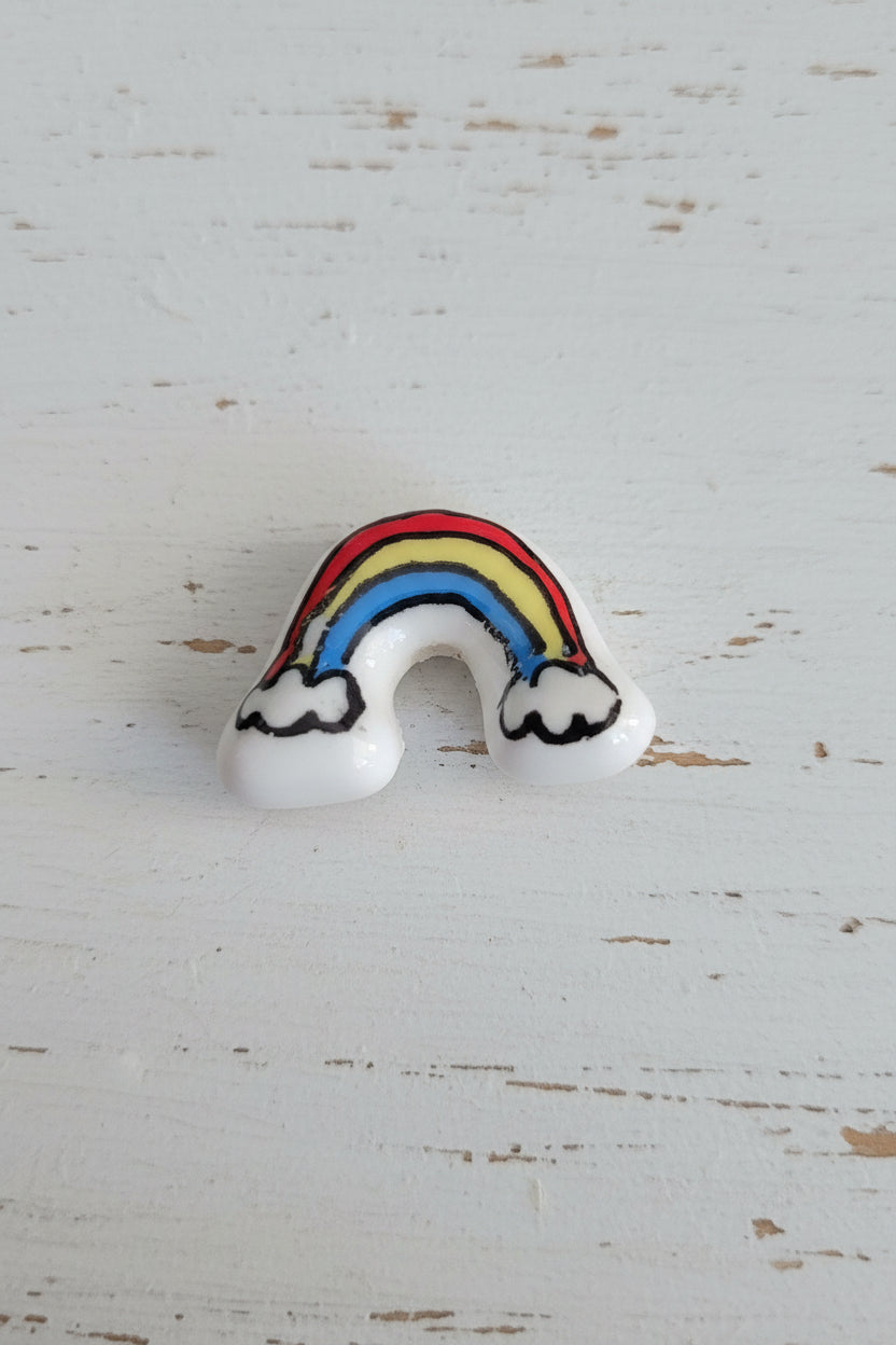 A small rainbow-shaped bead with white cloud ends. The rainbow has red, orange, yellow, blue, and black outlined details..