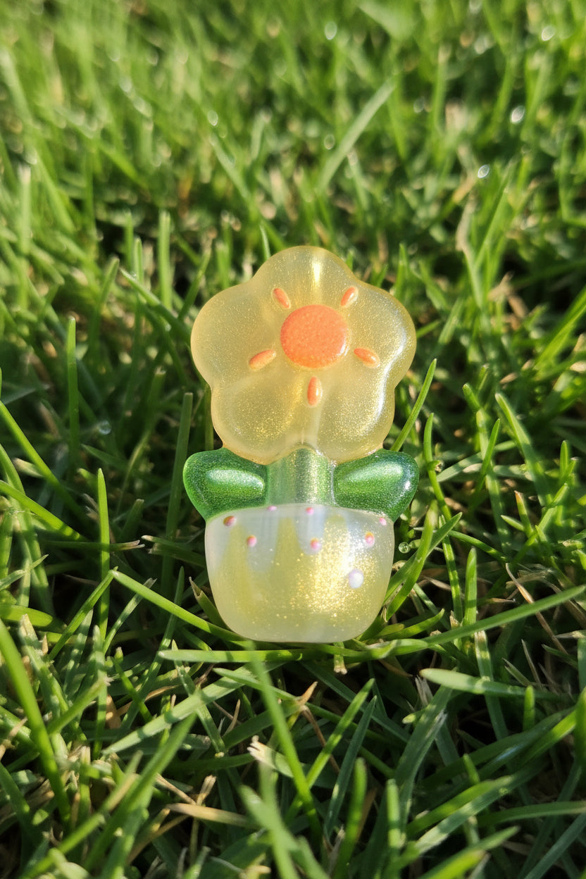 A small translucent flower-shaped bead, possibly a hair clip or charm, resting on green grass outdoors. The flower has a yellow-green translucent bloom with a bright orange center, green stems, and a sparkly pale yellow-green base.