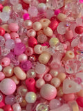 Assorted pink and pastel craft beads, including round glass crystal beads , faceted crystal beads, and cylindrical tubes, in shades of pink, red, and cream.