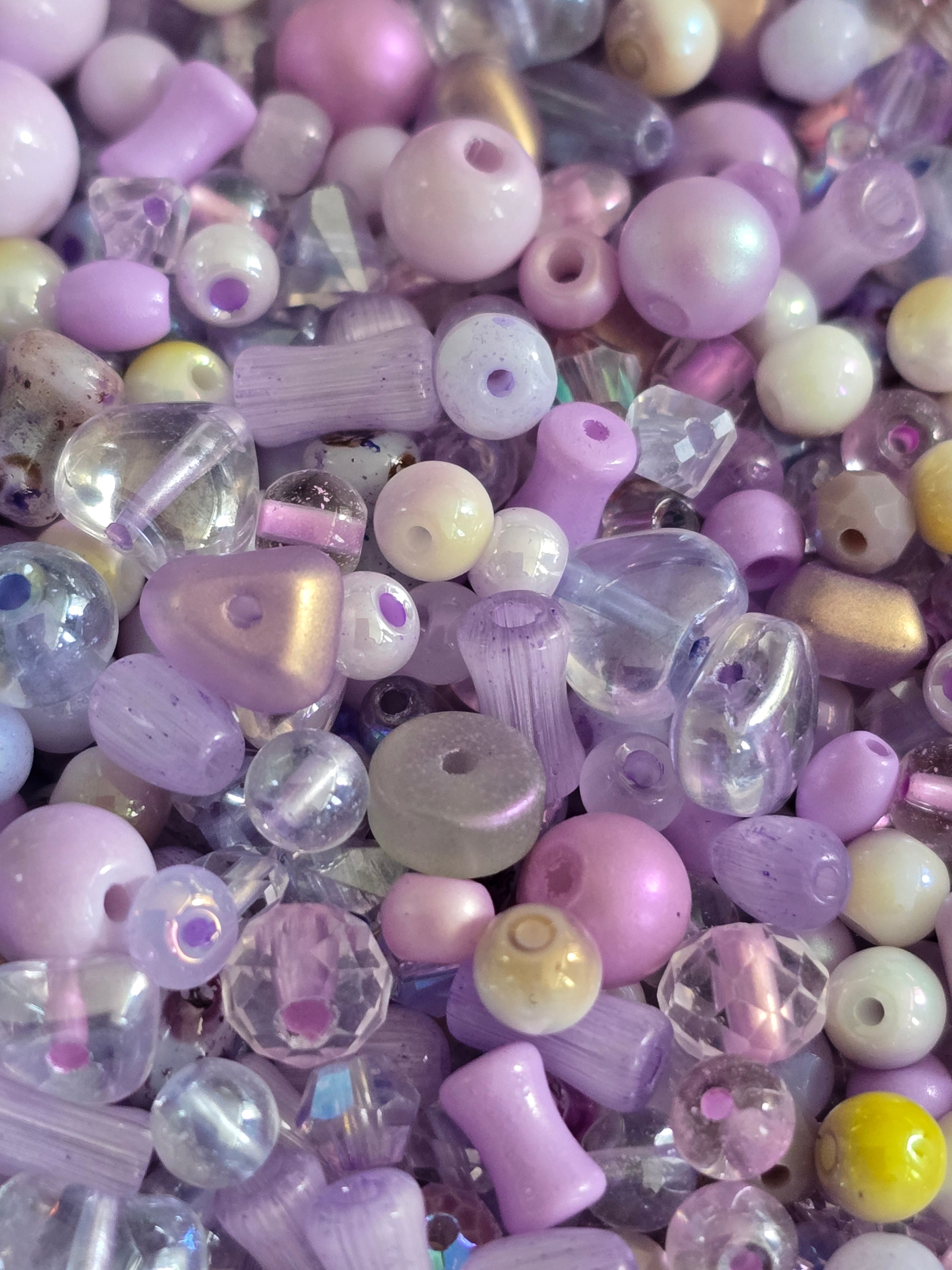 A dense pile of assorted craft beads in pastel lavender, lilac, pale pink, cream, and clear colors, showing various bead shapes and finishes.
