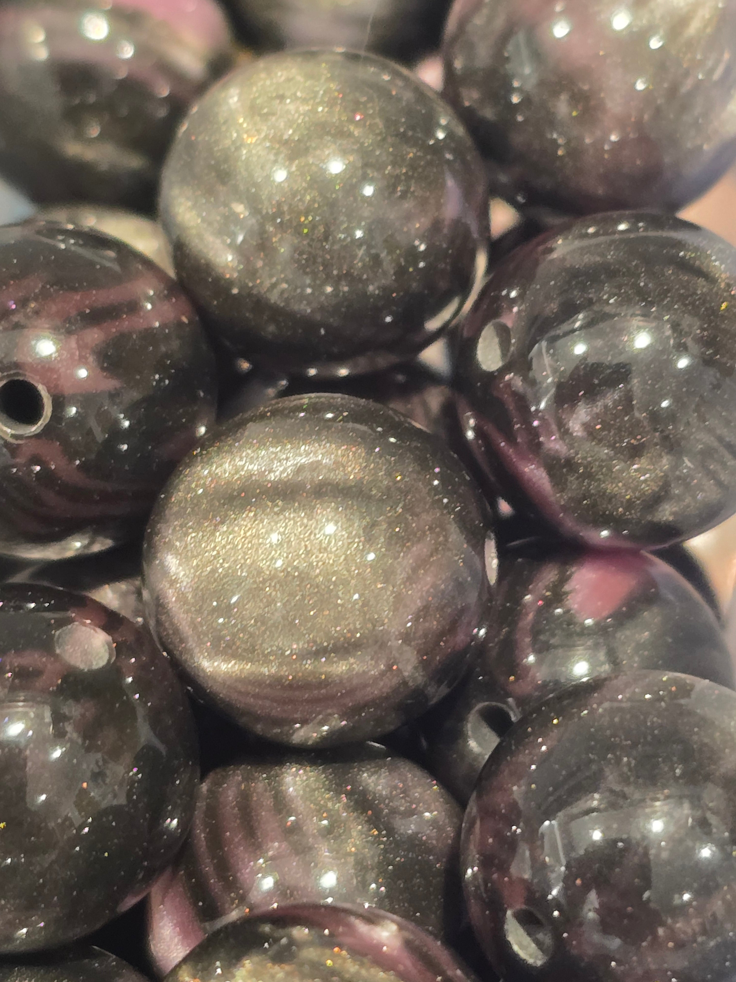 A close-up of glossy round beads with a smoky black and metallic gold glitter finish, showing a cat-eye effect under light.