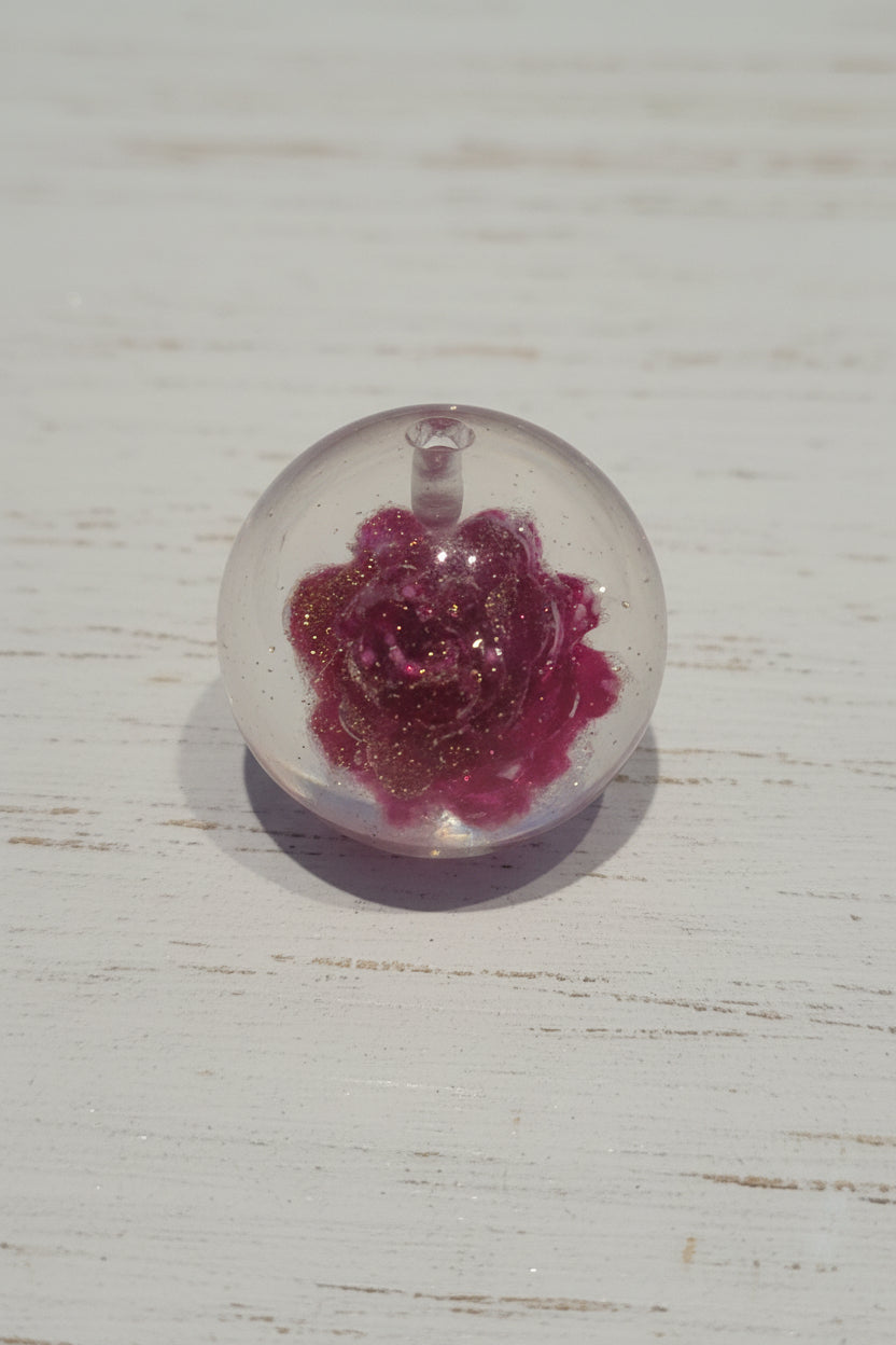 A clear round resin bead containing a pink glittery rose design with gold foil flakes.