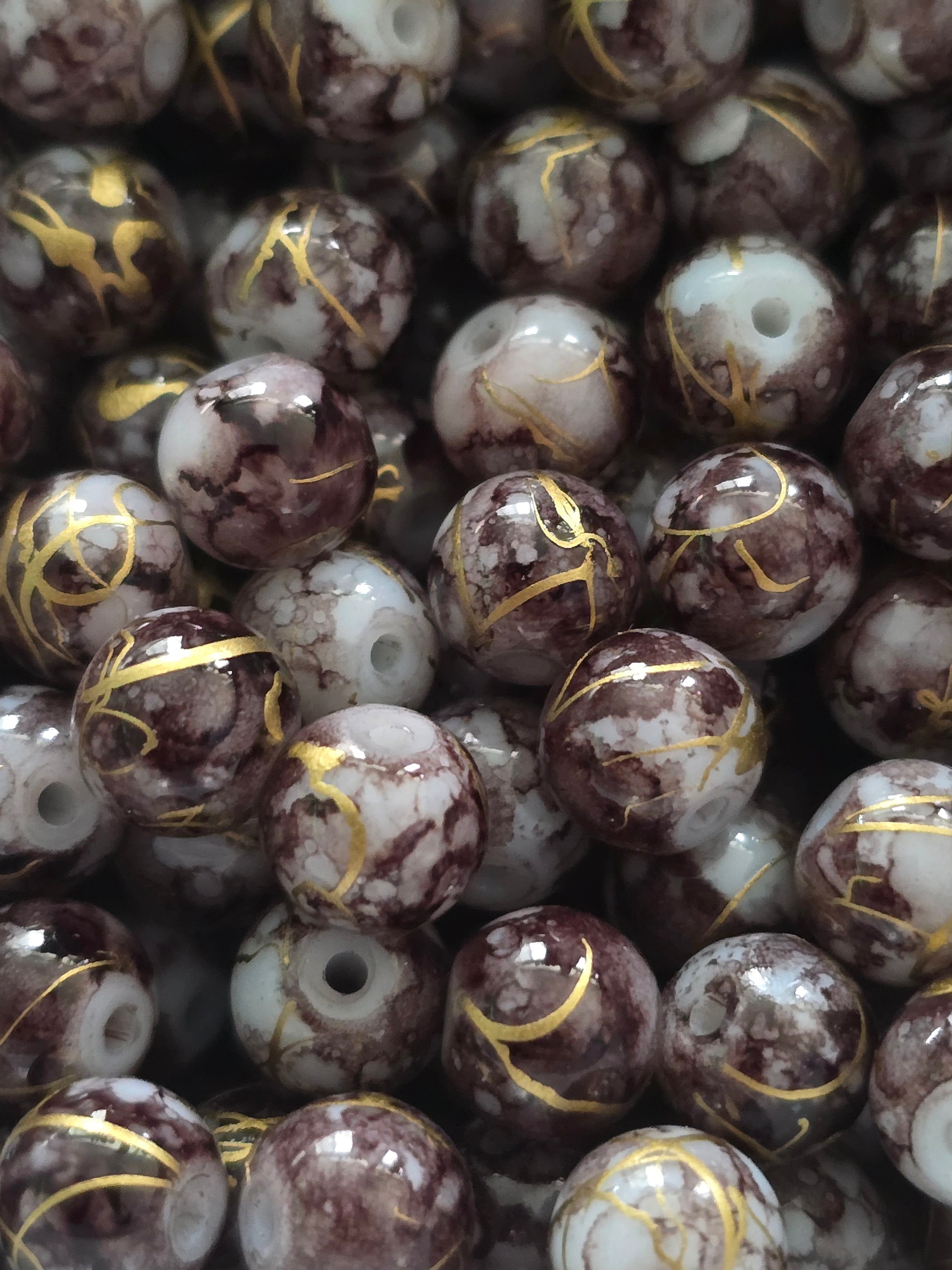 A close-up pile of round glass beads with a marbled pattern of dark brown, white, and black, accented with thin metallic gold veins running through the center.