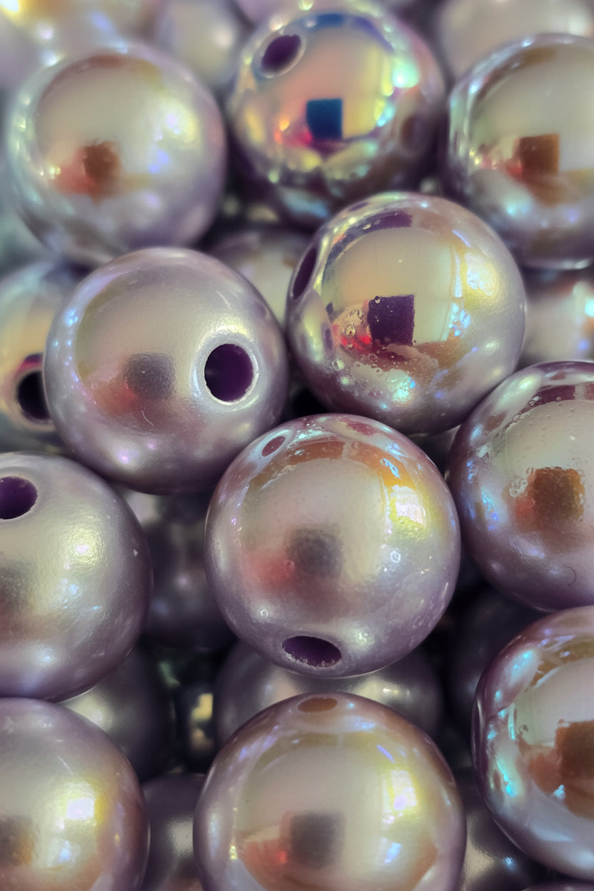 A close-up pile of round, iridescent pearl-like beads with a metallic lavender-gray sheen and small holes, reflecting light.