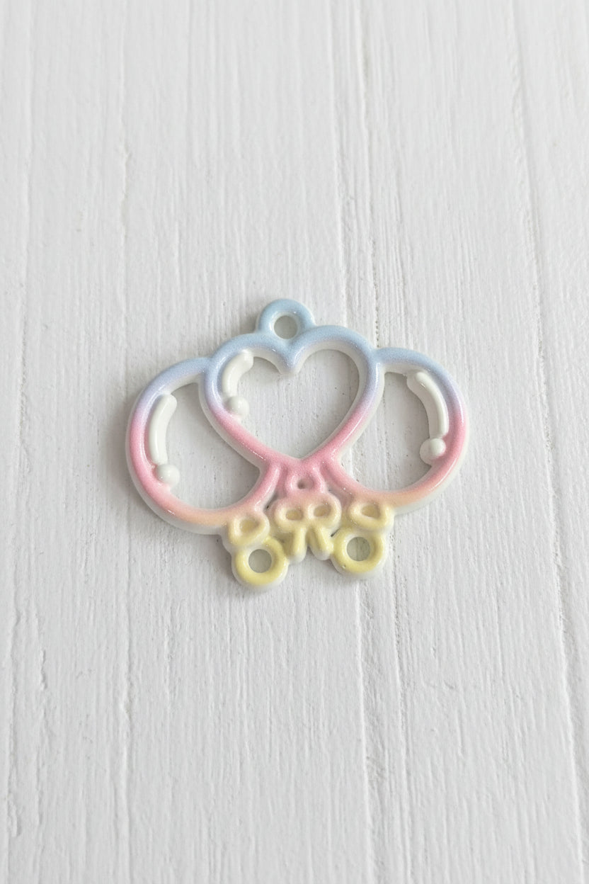 A small decorative metal charm with a pastel rainbow gradient, featuring a heart and crescent moon design with three hanging loops.