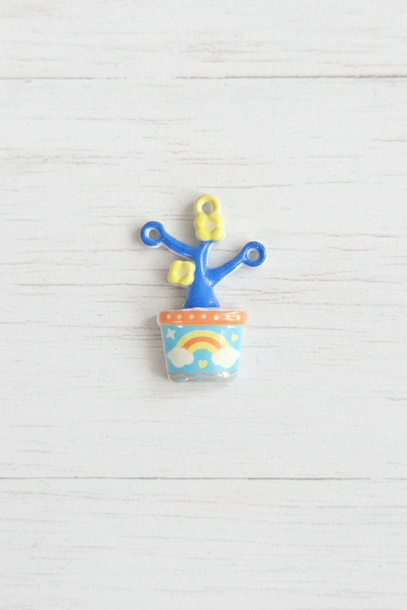 A small decorative pendant f metal featuring a blue anchor-like figure with yellow loops and a cylindrical container decorated with a rainbow and clouds.