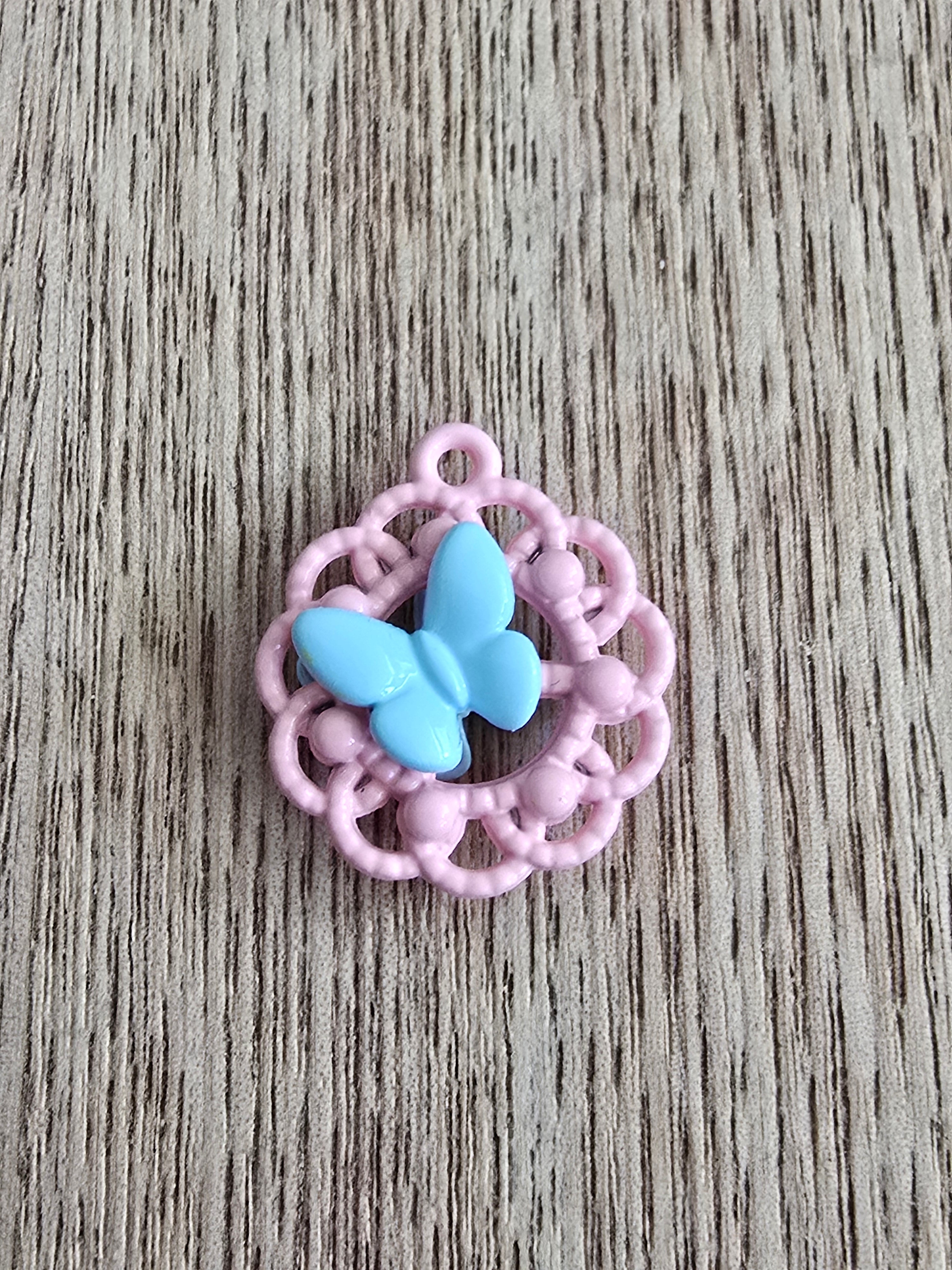 A small metal pendant featuring a light blue butterfly motif surrounded by a pink filigree-style circular border.