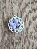 A small white metal circular lace-like pendant with a central lavender butterfly.