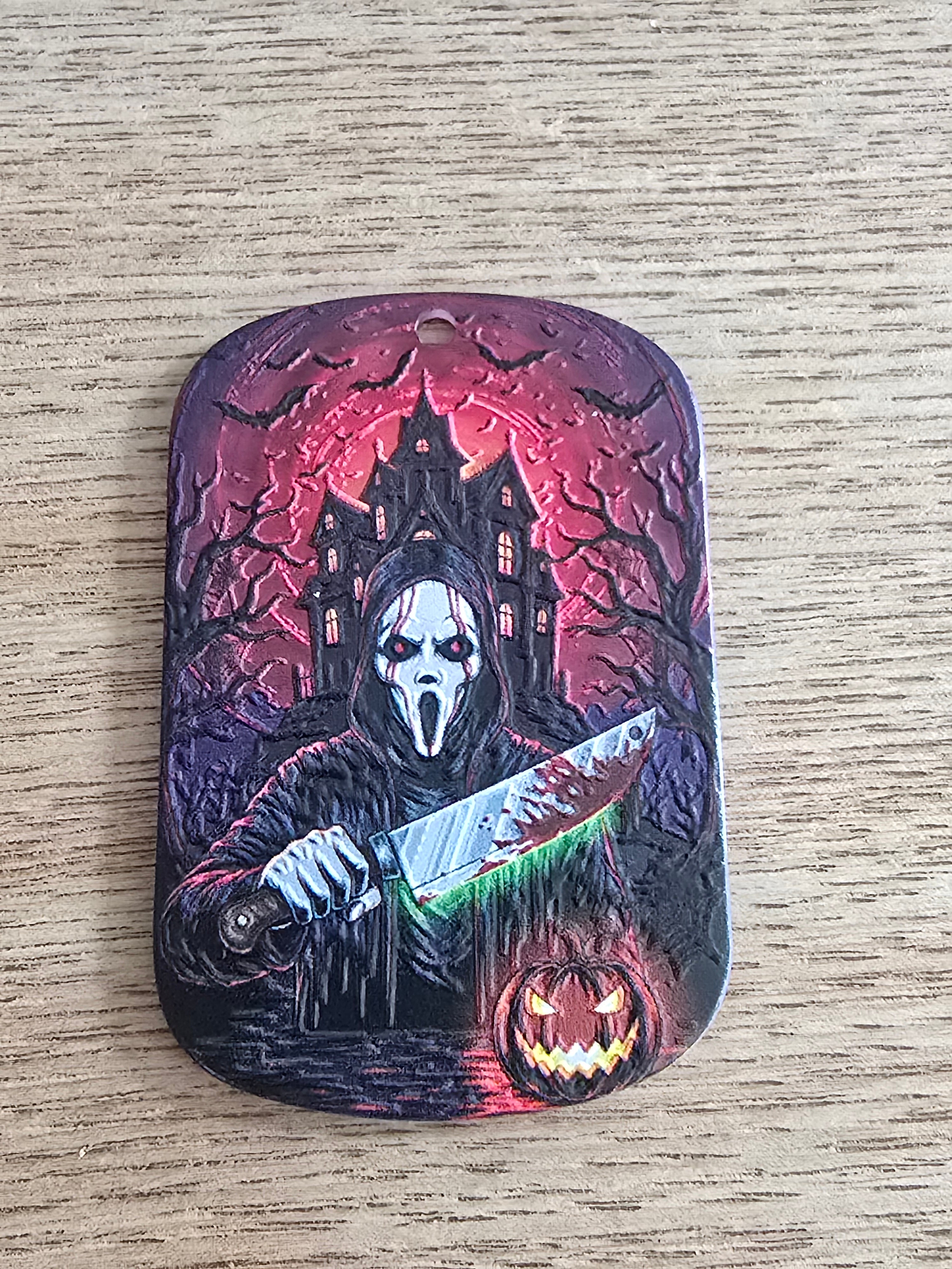 A rectangular acrylic tag with rounded corners, featuring a detailed Halloween-themed illustration of a ghost face, a knife, a haunted house, and a jack-o'-lantern on a textured dark background.