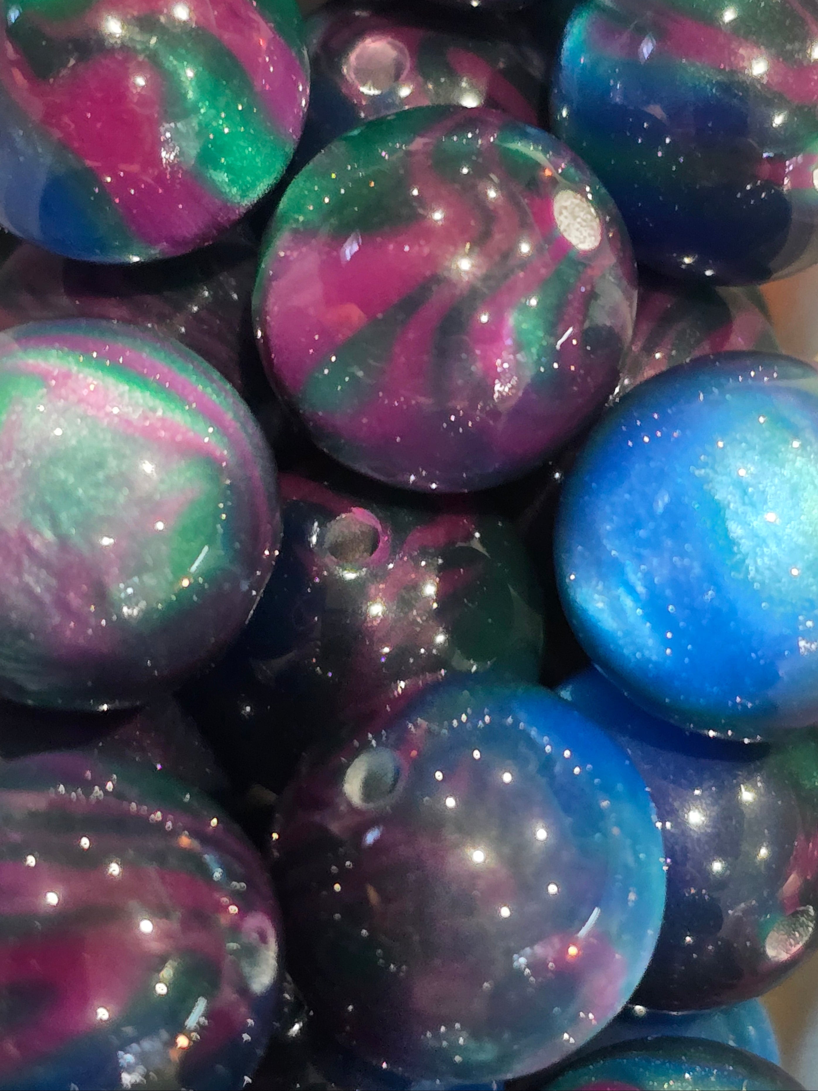A close-up pile of round, glossy beads with a swirling galaxy pattern in blue, purple, green, and pink, filled with glitter.