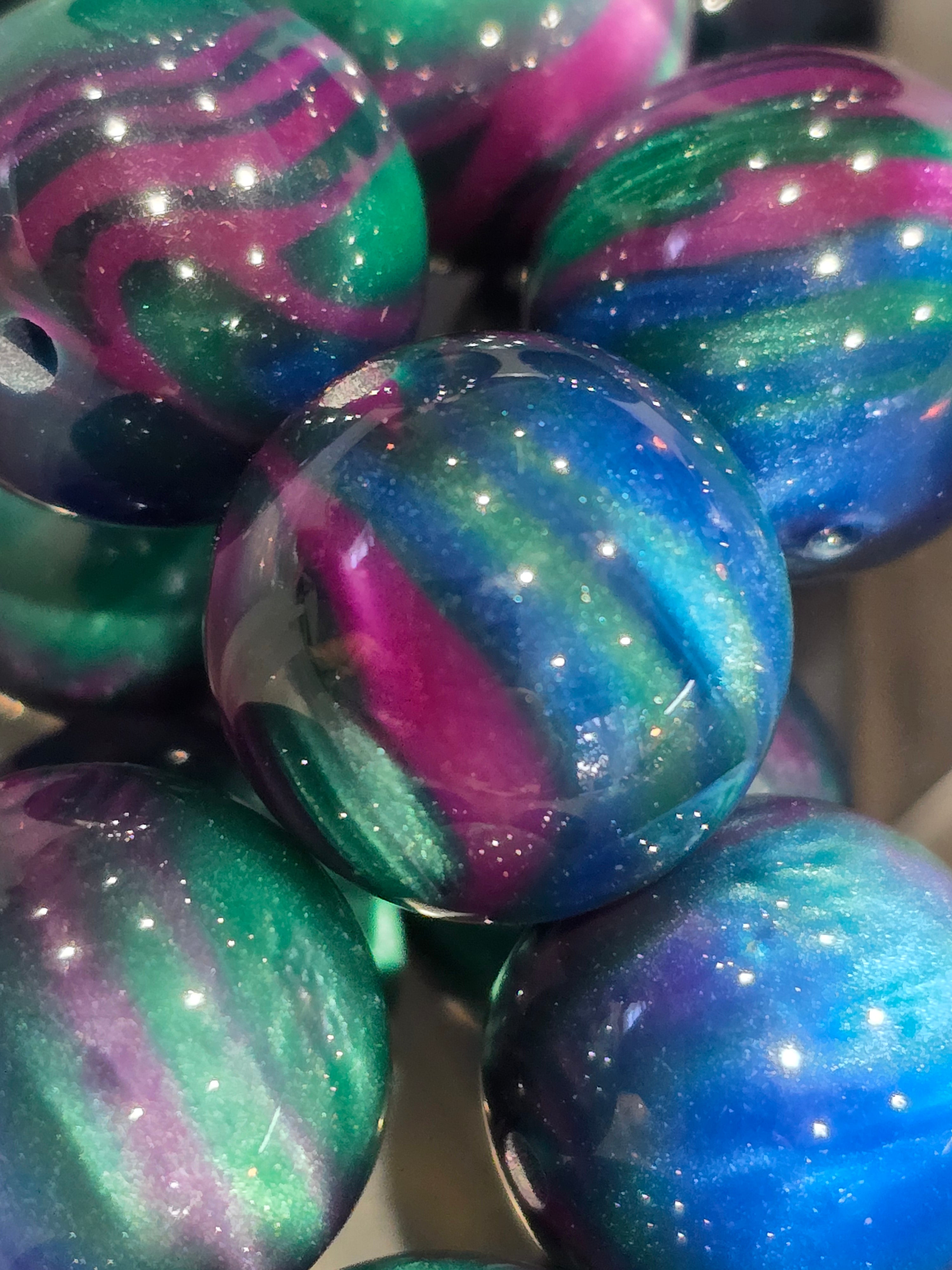A close-up of glossy, round 20mm resin beads with a marbled cosmic pattern in blue, green, and purple, featuring a subtle glitter finish.