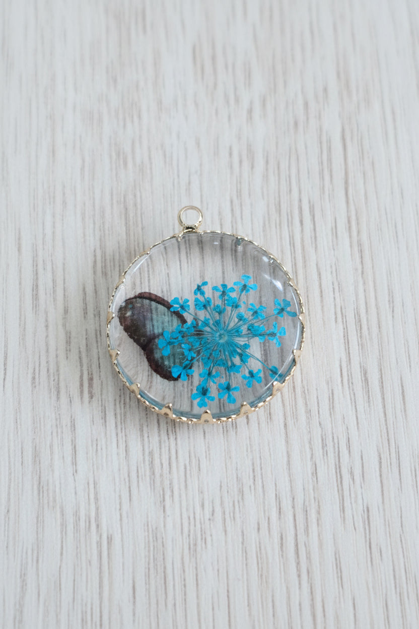 A round resin pendant featuring a dried blue flower arrangement inside, set in a gold-toned scalloped bezel with a small hanging loop