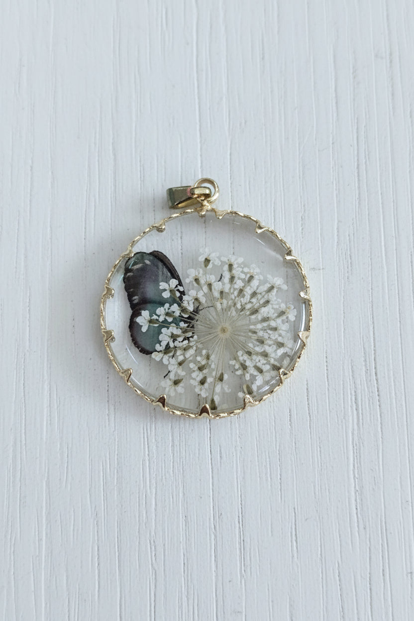 A round glass cabochon pendant featuring a white dandelion and a small dark butterfly, set in a gold-toned metal frame.