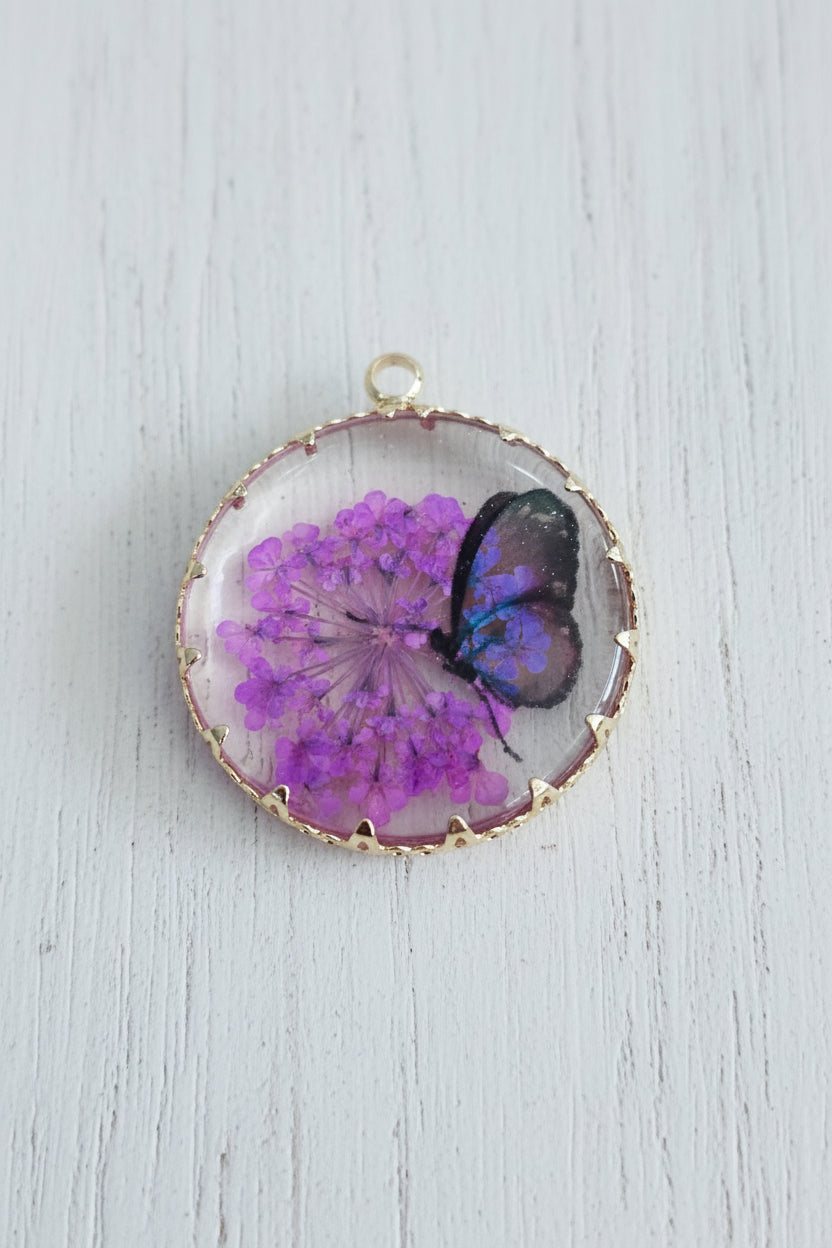 A round pendant or charm with a - gold toned scalloped border, featuring a preserved purple flower cluster and a black butterfly inside a clear resin dome.