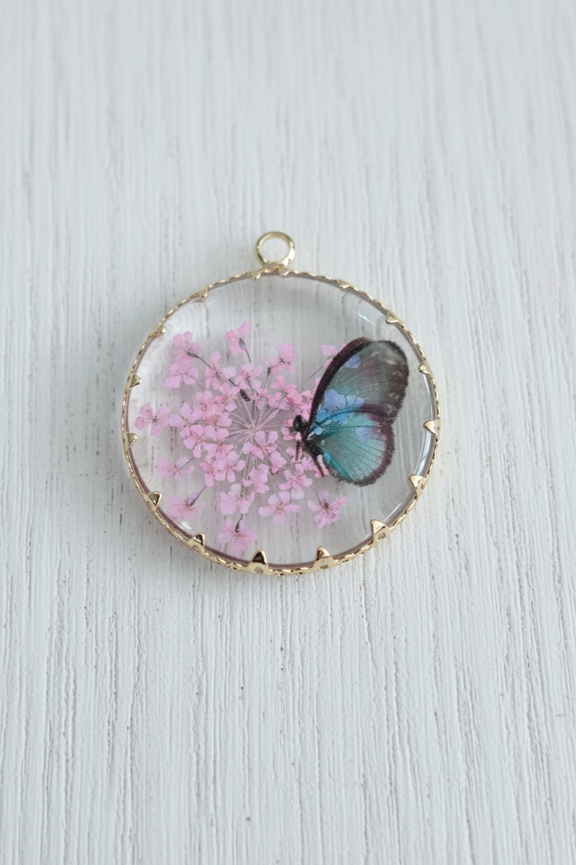 A round glass pendant charm with a gold-toned rim, featuring a preserved blue butterfly and pink flowers inside.