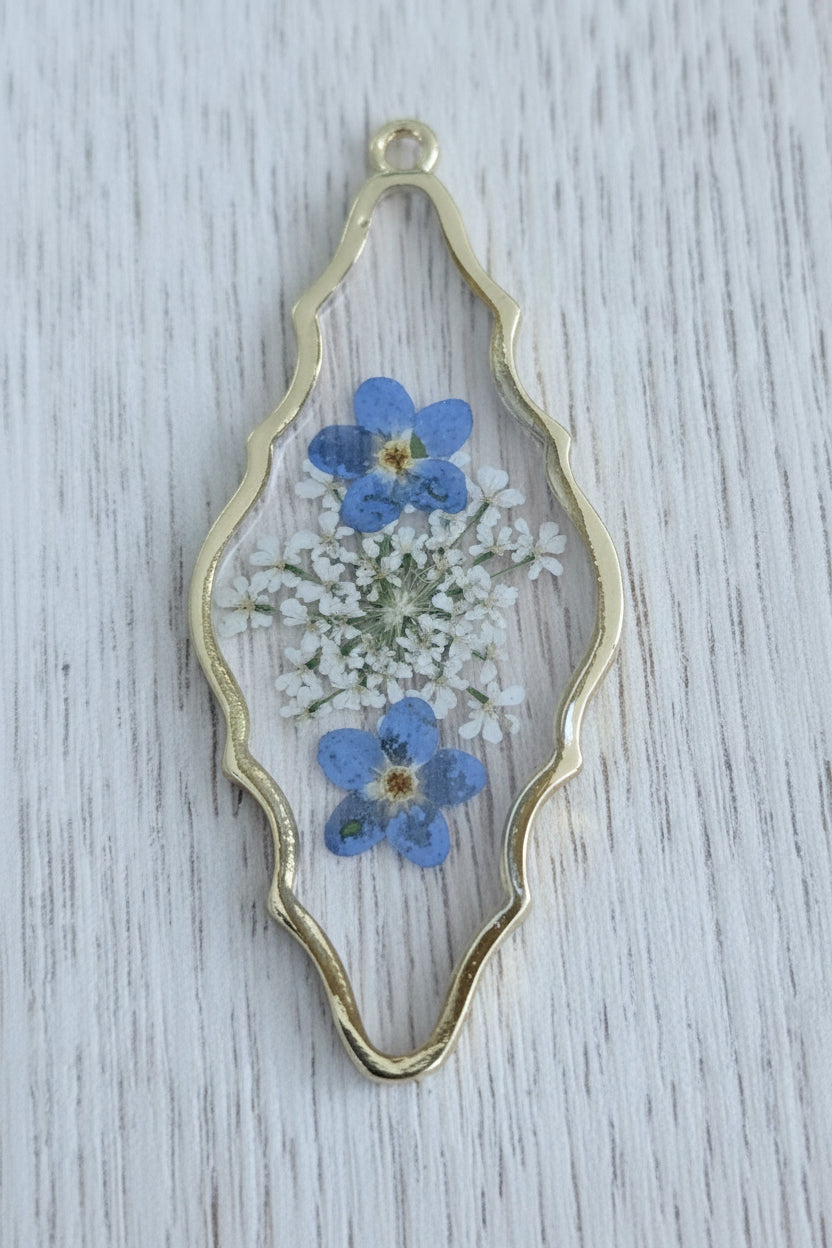 A gold-tone metal pendant containing two blue forget-me-not flowers and white yarrow.