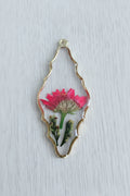 A decorative pendant with an irregular wavy outline, featuring a gold-toned metal border containing a pressed pink flower and green botanical elements.
