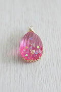 A pink teardrop-shaped pendant with a textured, ribbed surface containing iridescent glitter, finished with a gold-tone bail.