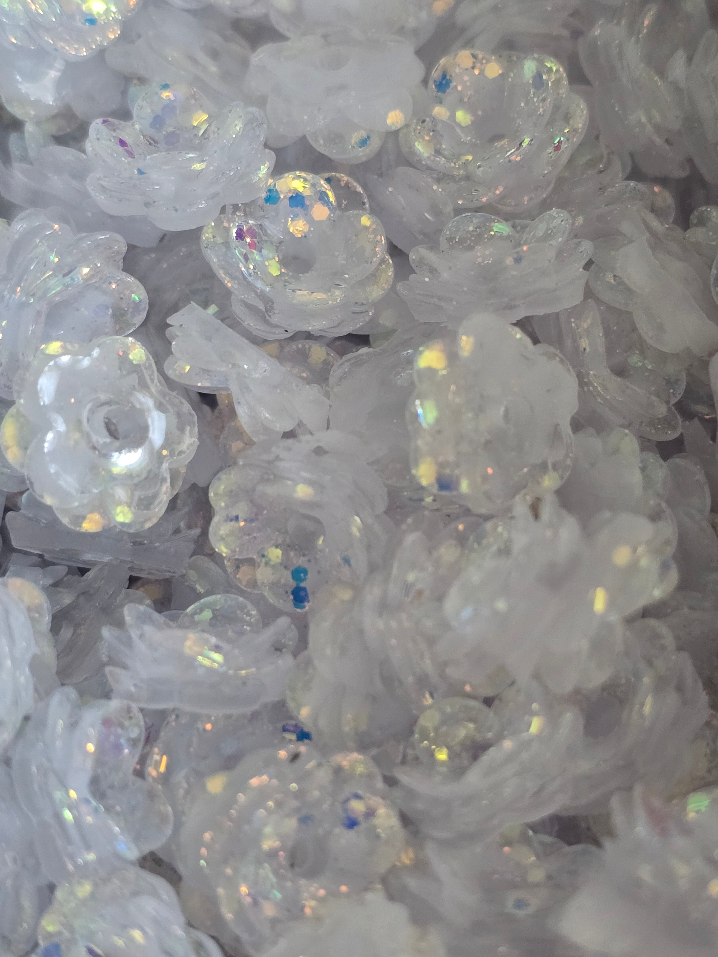 A close-up pile of clear, flower-shaped beads with iridescent glitter and small blue sequins.