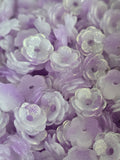 A close-up pile of small lavender and translucent white flower-shaped beads with glittery highlights.