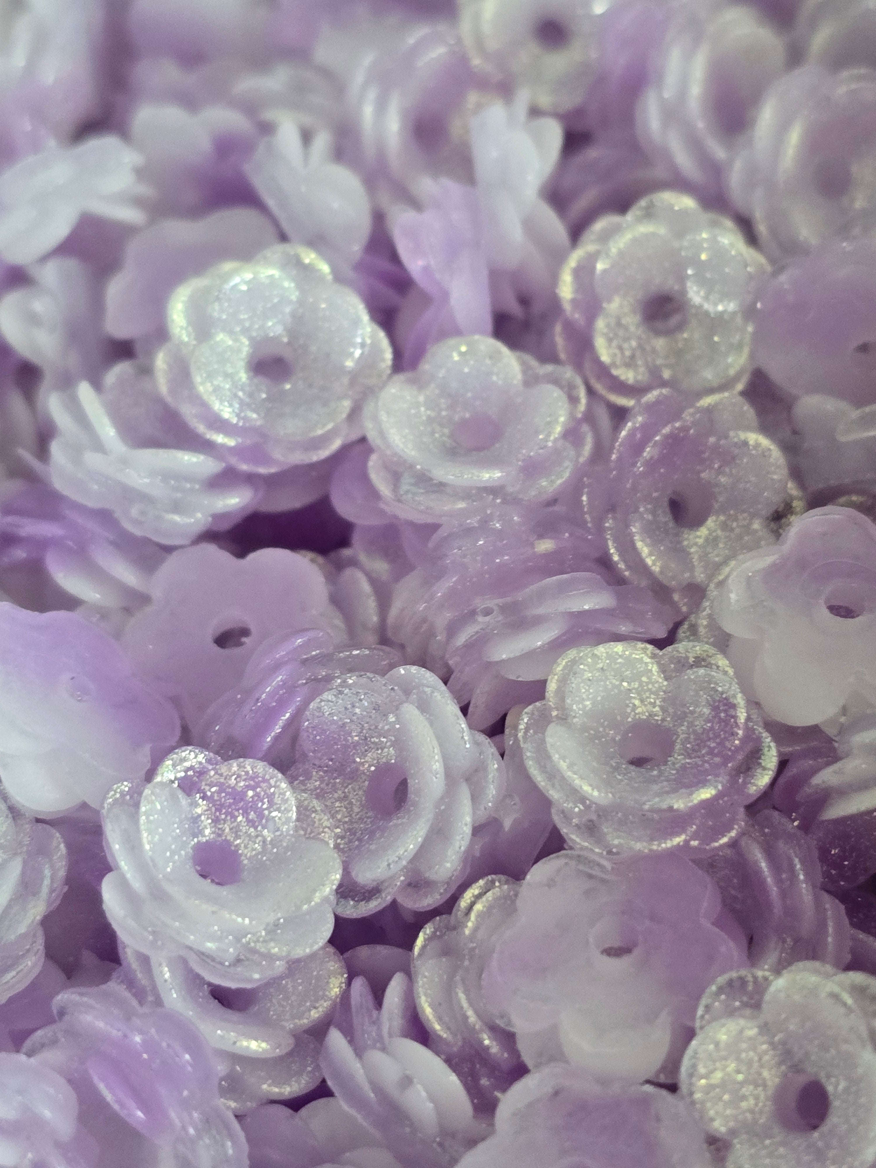 A close-up pile of small lavender and translucent white flower-shaped beads with glittery highlights.