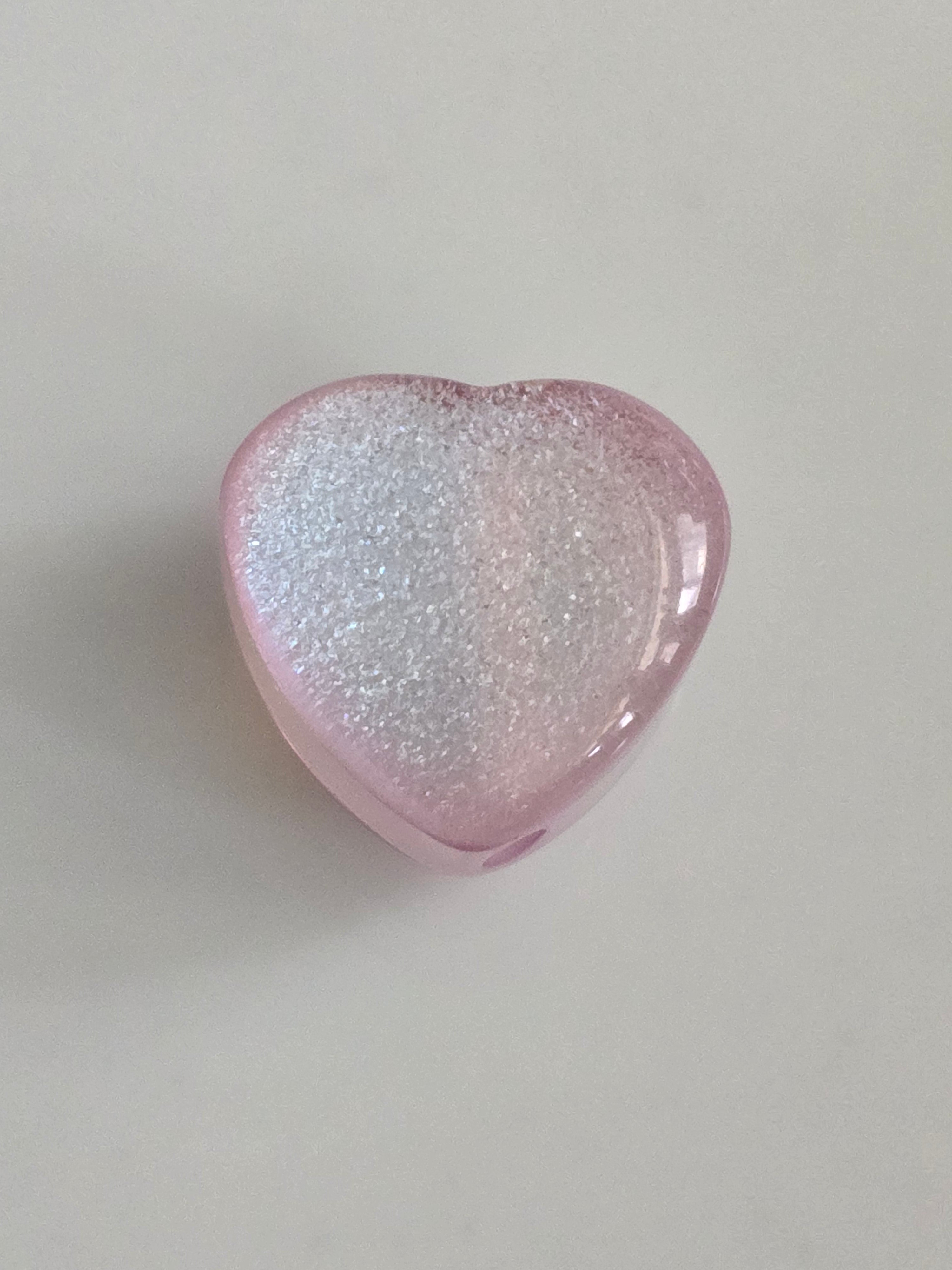 A heart-shaped pink resin bead with shimmering silver glitter, shown on a neutral light gray background.