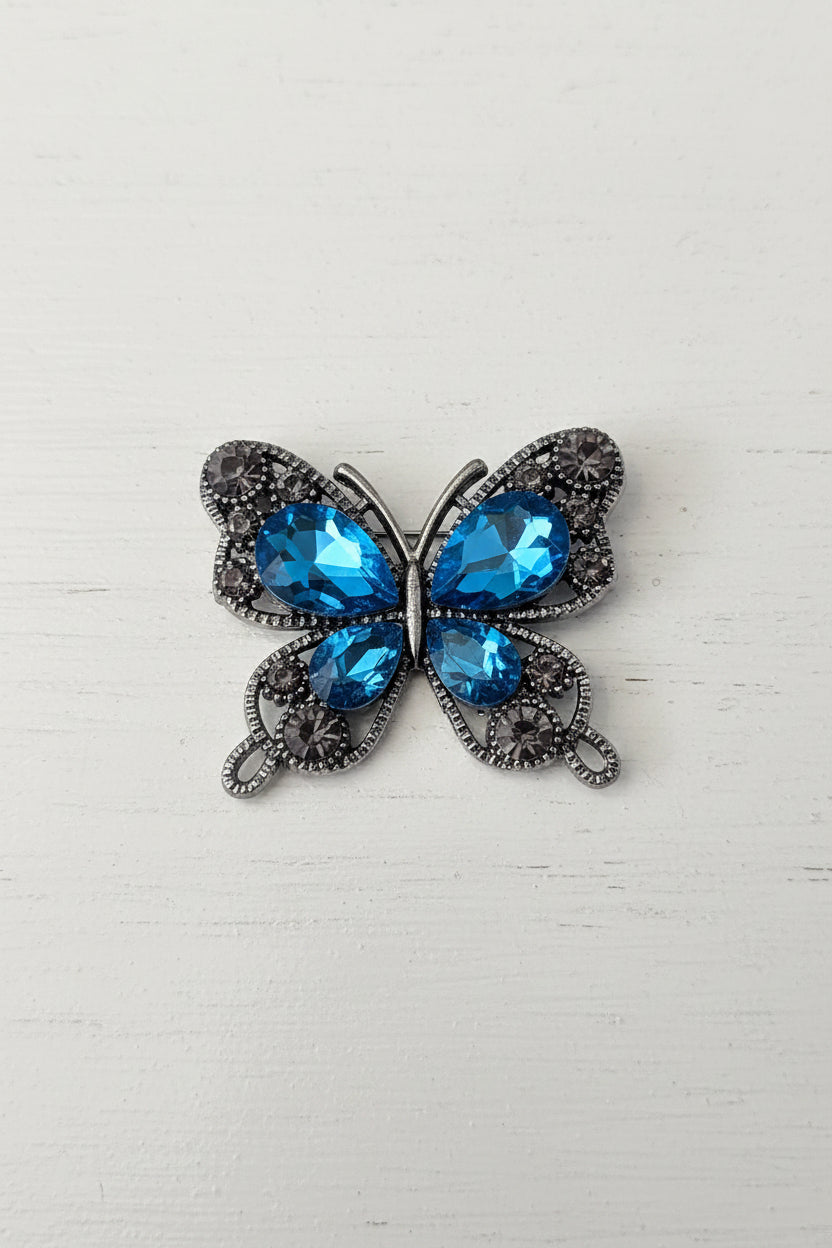 A butterfly-shaped flatback featuring large faceted blue rhinestones in the centre and smaller blue and smoky grey rhinestones set in a silver-tone.