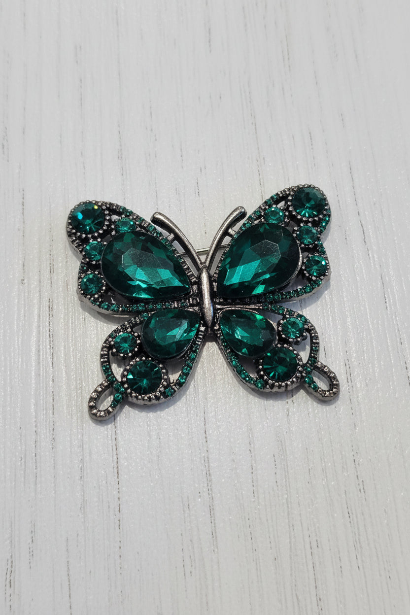 A butterfly-shaped flatback featuring large faceted emerald green rhinestones in the centre, surrounded by silver-toned filigree and small clear rhinestones.