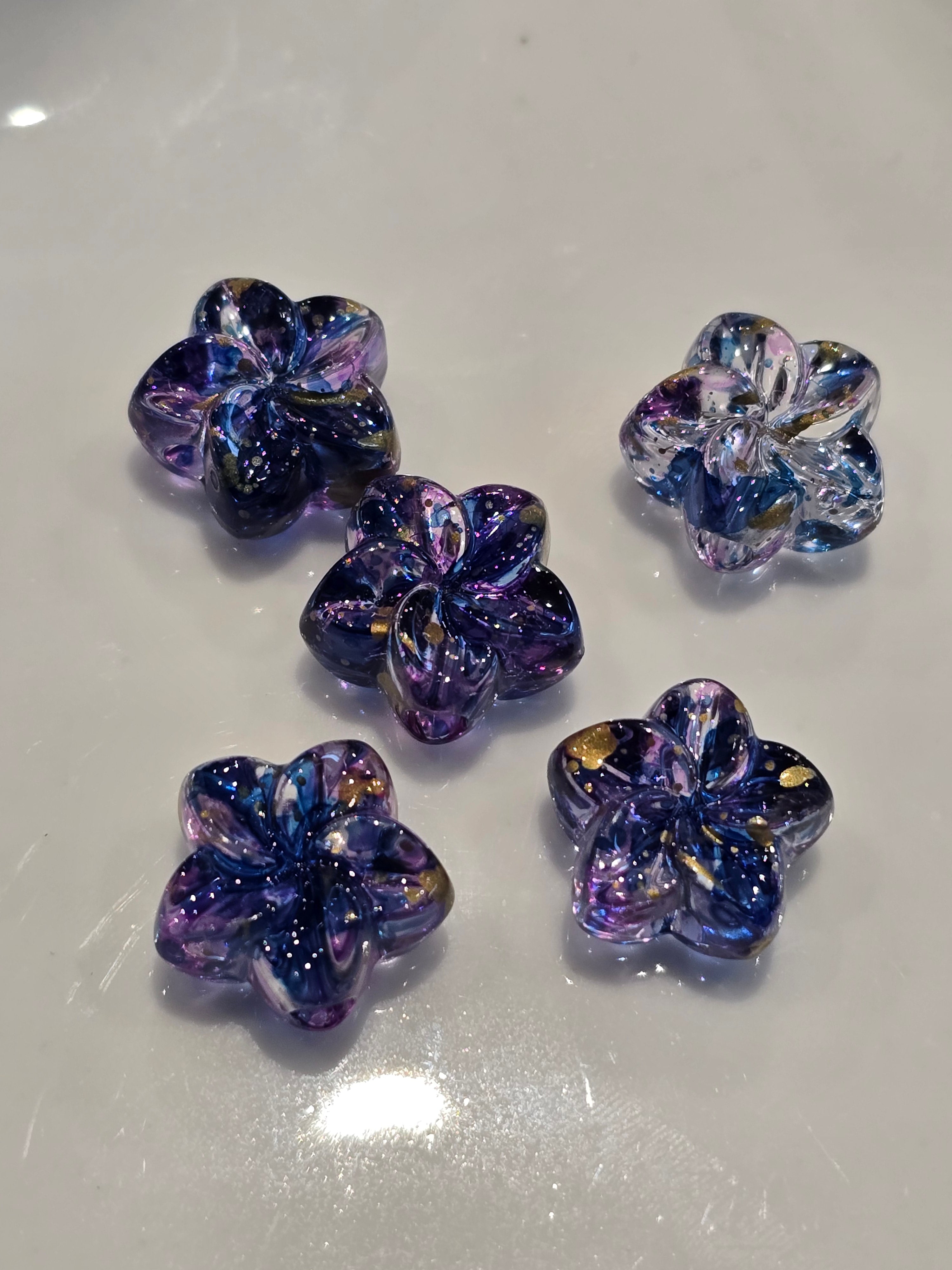 Acrylic flower beads with a swirling iridescent pattern of purple, blue, and gold, displayed on a glossy white surface.