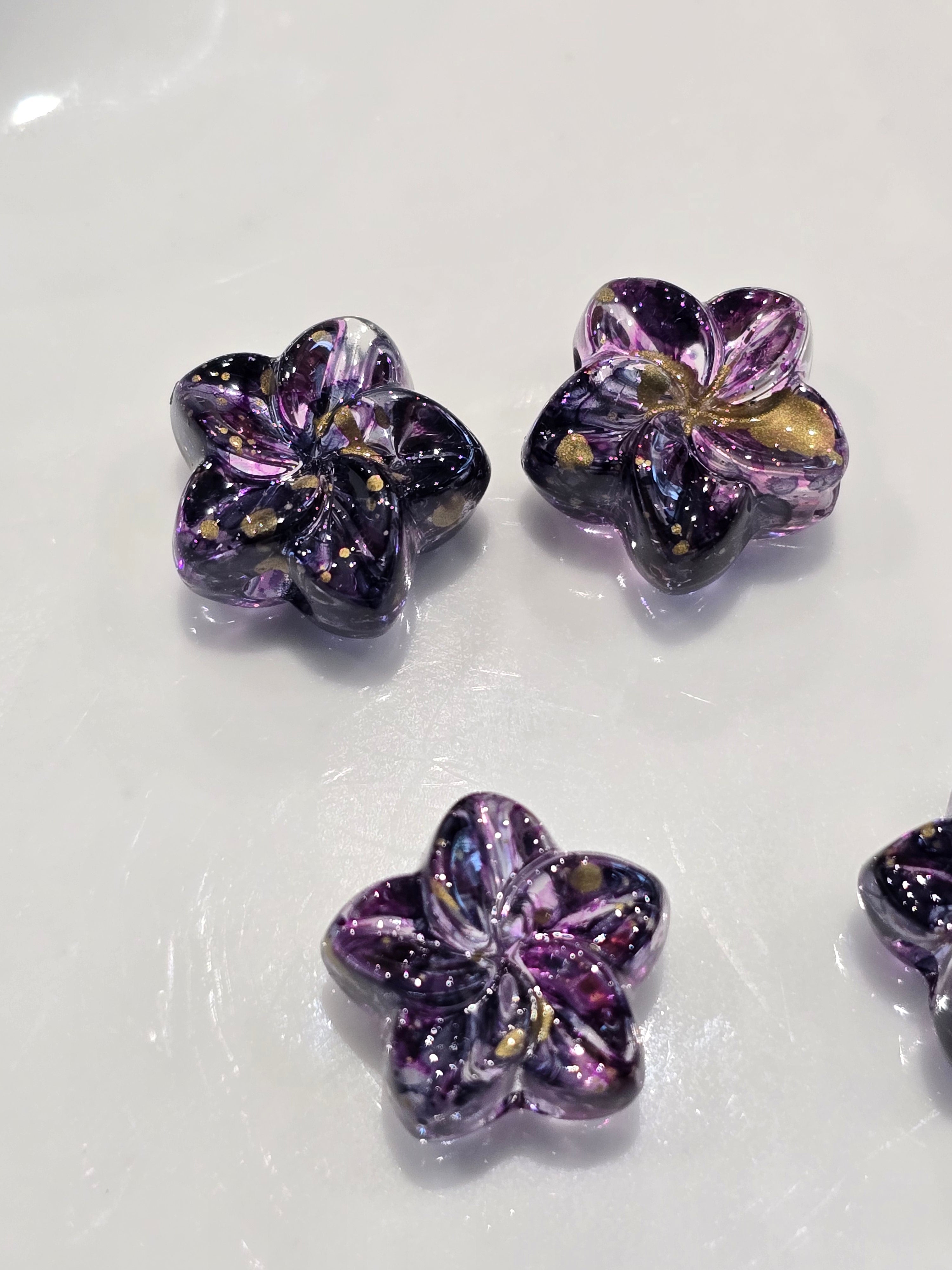 Acrylic flower-shaped beads with a purple and black galaxy pattern and gold glitter, displayed on a glossy white surface.