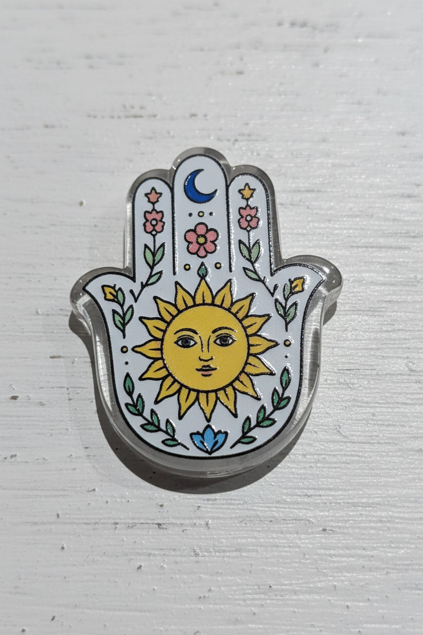A Hamsa hand acrylic bead with a silver-toned metal border and a white enamel background, decorated with a yellow sun face, green leaves, pink flowers, and a blue crescent moon.