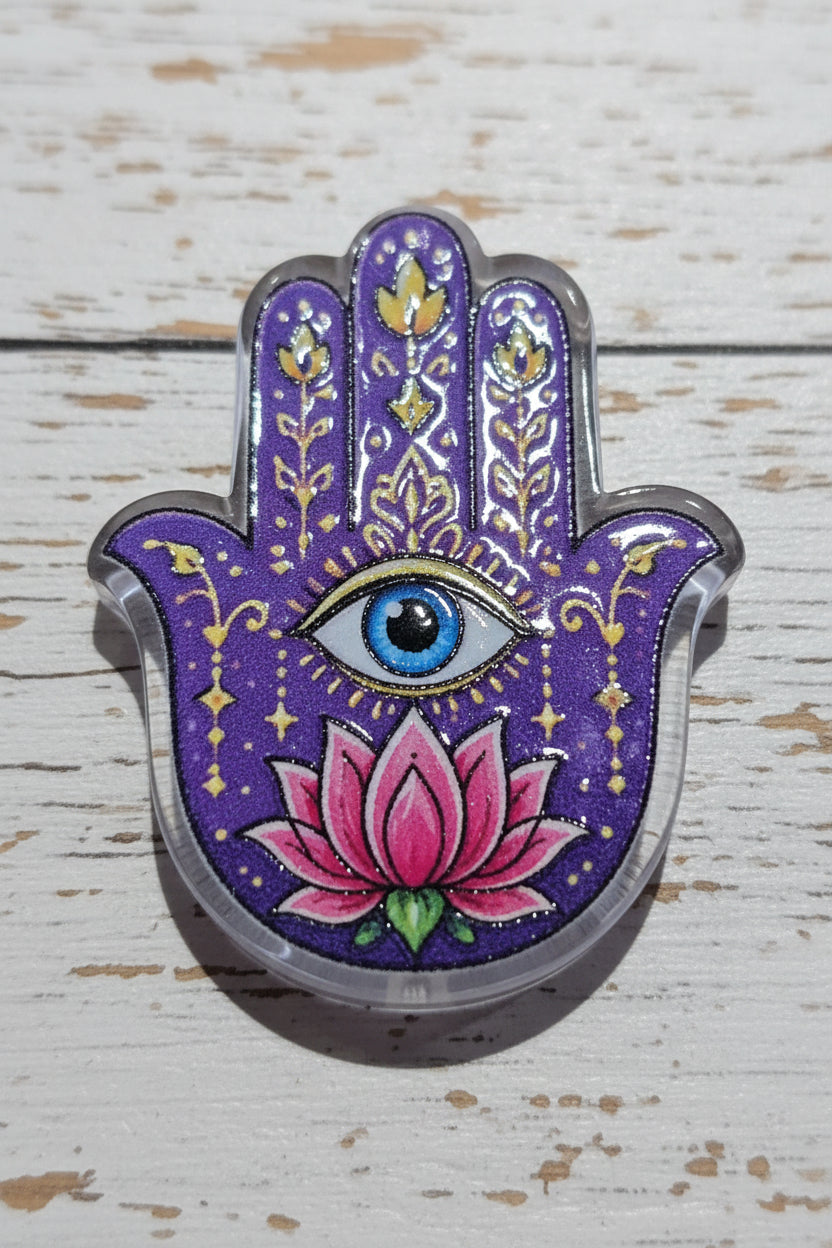 A Hamsa hand acrylic bead featuring a purple glitter background with gold filigree patterns, a central blue and black eye, and a pink lotus flower with green leaves at the bottom.