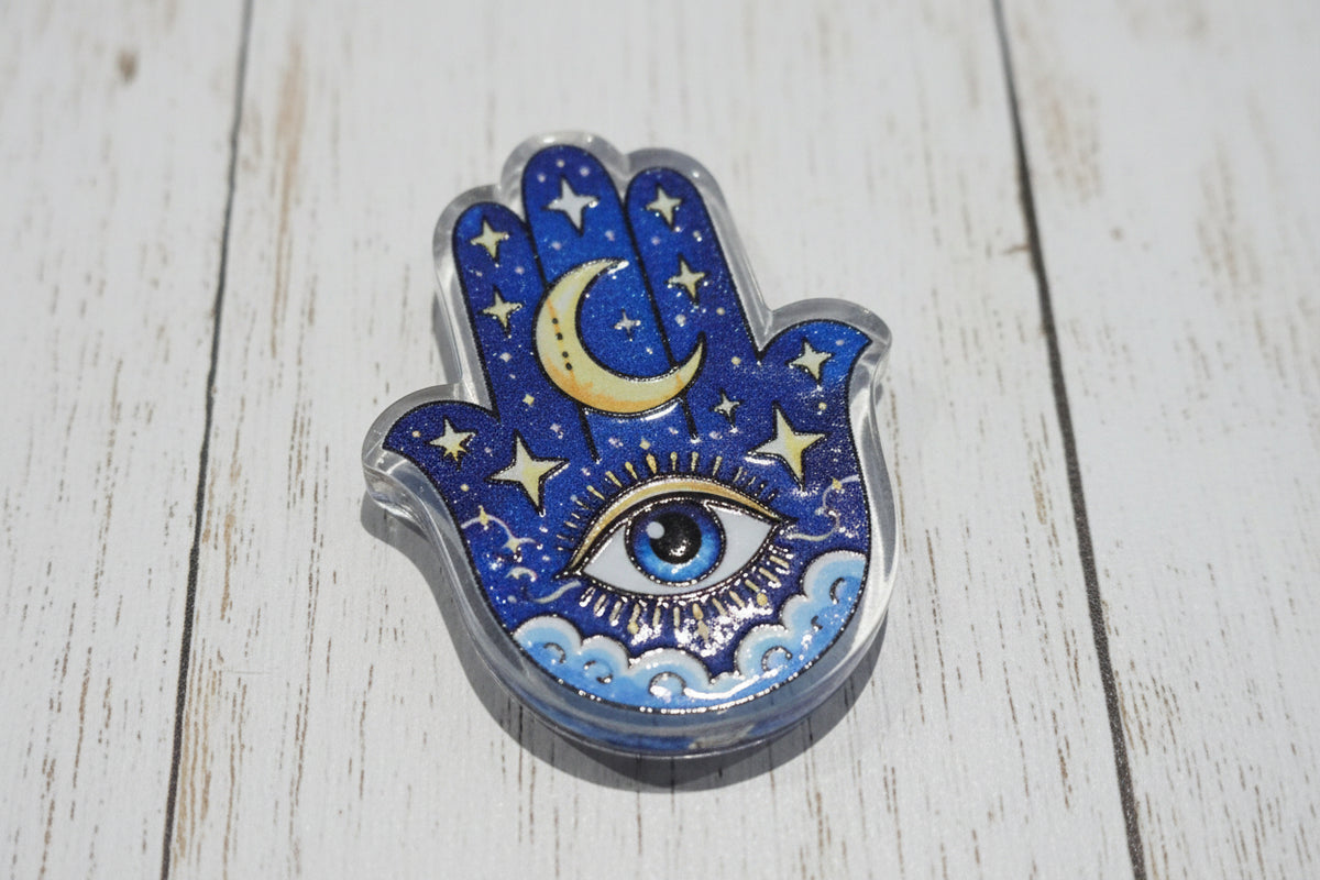 A Hamsa hand acrylic bead featuring a celestial design with a blue glitter background, gold crescent moon, stars, and an all-seeing eye.