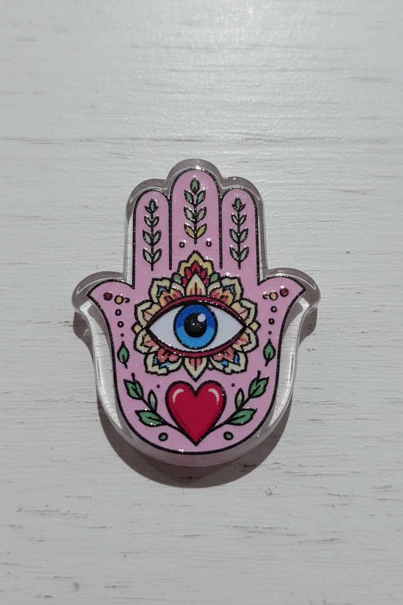 A pink Hamsa acrylic bead with a glittery silver border, featuring a blue and white eye, green leaves, a red heart, and floral patterns.
