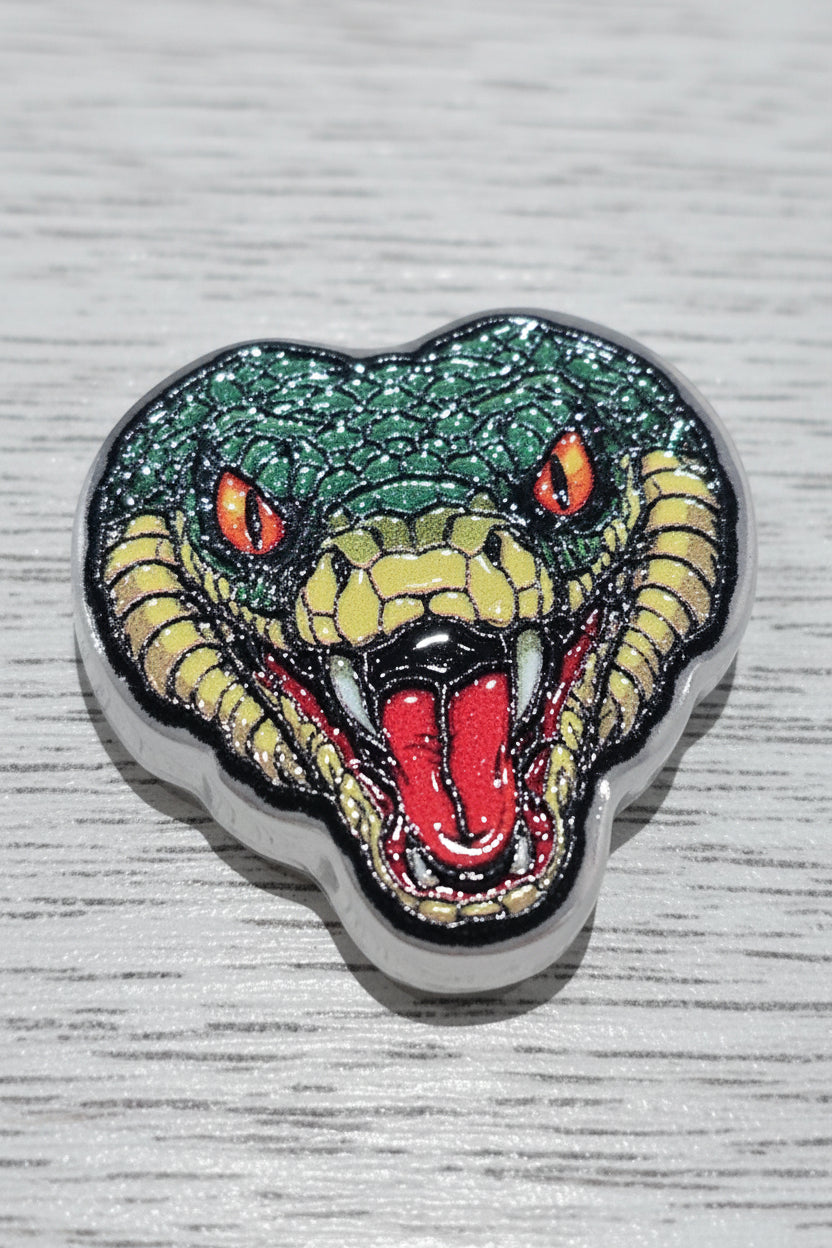 A heart-shaped acrylic bead featuring a green and yellow snake head with red eyes, a red tongue, and black outlines.