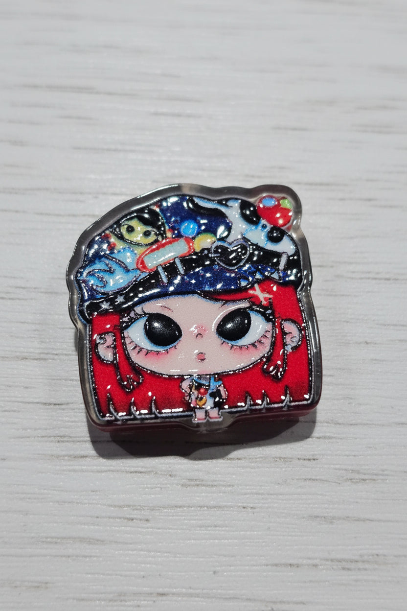 A small acrylic bead featuring a stylized cartoon face with large black eyes, a red lower face and hair, and a dark blue glittery hat .