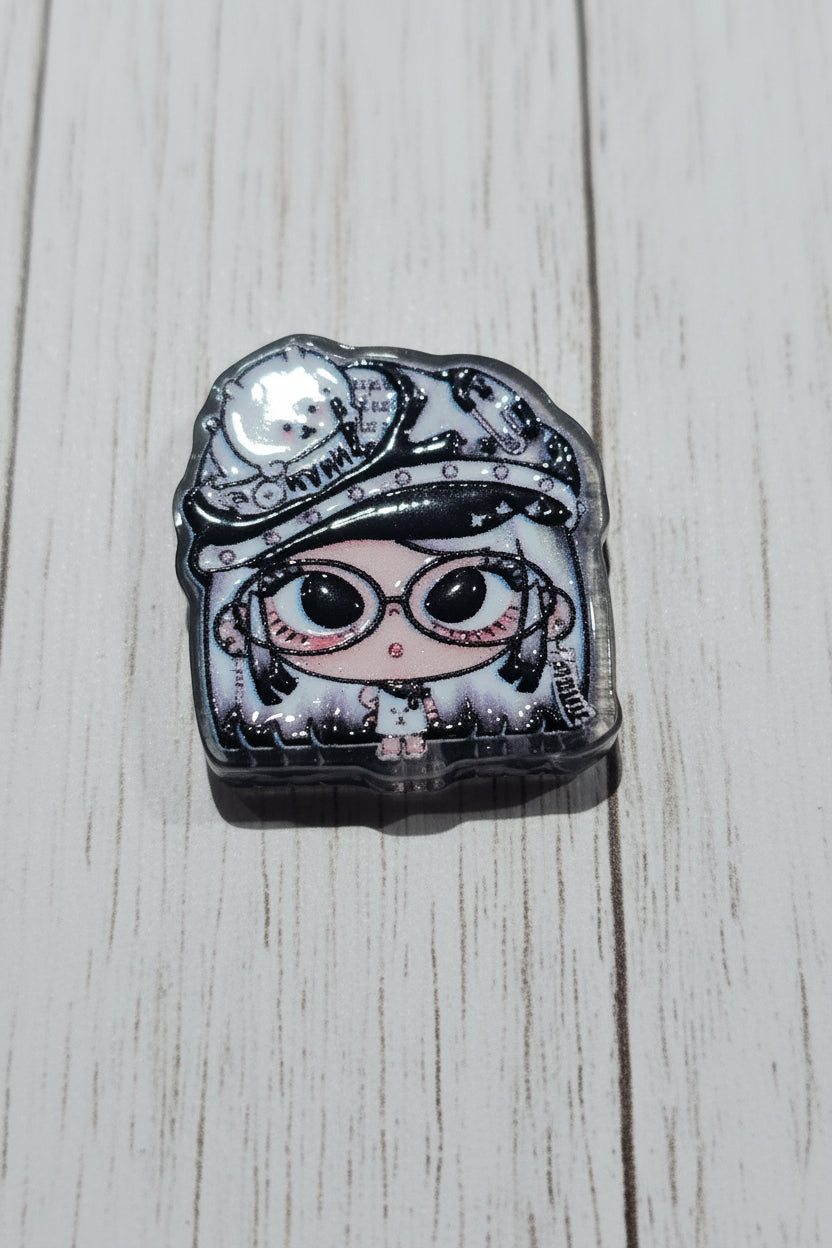 A small illustrated bead with a cartoon face wearing a hat and goggles, shown on a wood surface. The design is mostly black, white, and gray with pink accents around the eyes and mouth.