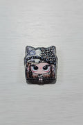 A square acrylic bead featuring a stylized anime doll face with large black eyes, a leopard-print hat, and glittery details.
